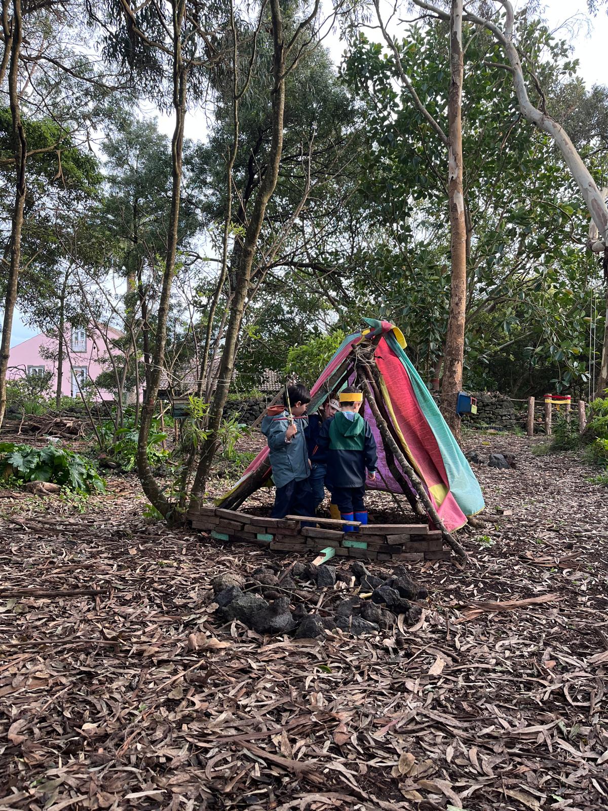 Crianças a brincar dentro de uma tenda construída com paus e tecido colorido